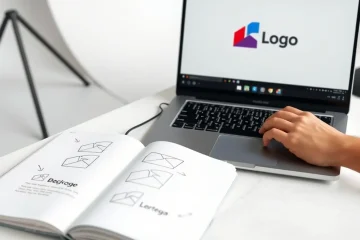 alt_text: A designer's hands refine a geometric logo on a laptop, with an open sketchbook showing early concepts in a bright, minimalist studio.